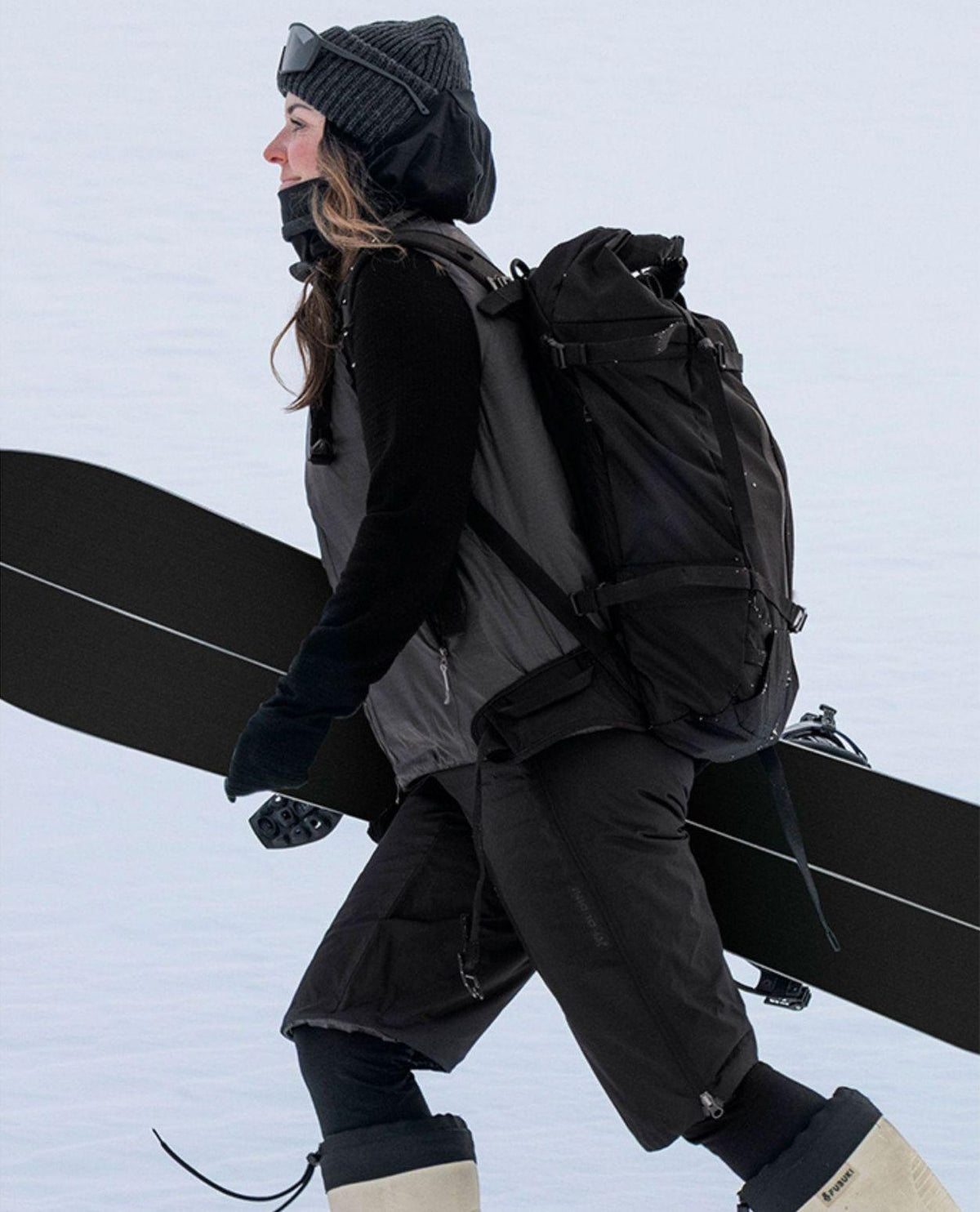 Kvinna i svart mössa, ryggsäck och svarta dunfria shorts som bär skidor utomhus i snö.