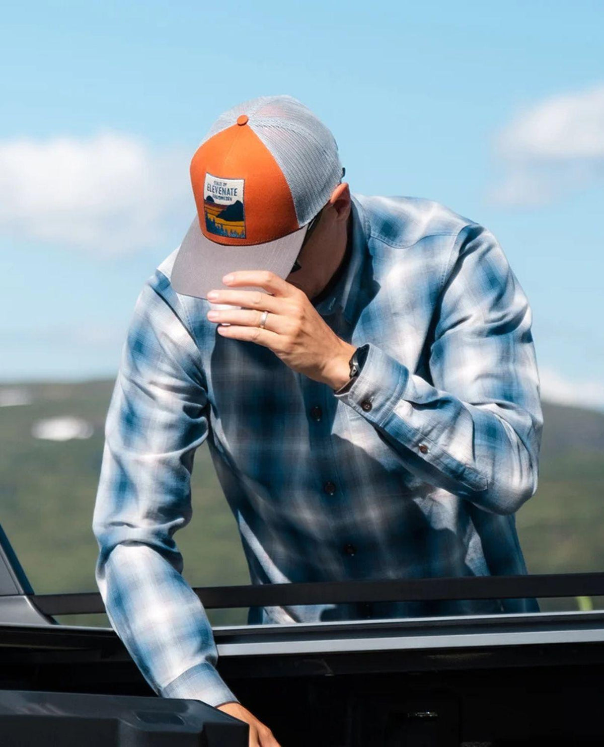 Person i en blårutig skjorta bär Elevenate Tofino Cap i färgen copper utomhus i ett naturlandskap.