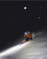 Skidåkare i orange jacka åker djupsnö på kvällen med Black Diamond Helio Carbon 108 topptursskidor under pannlampa i mörker.