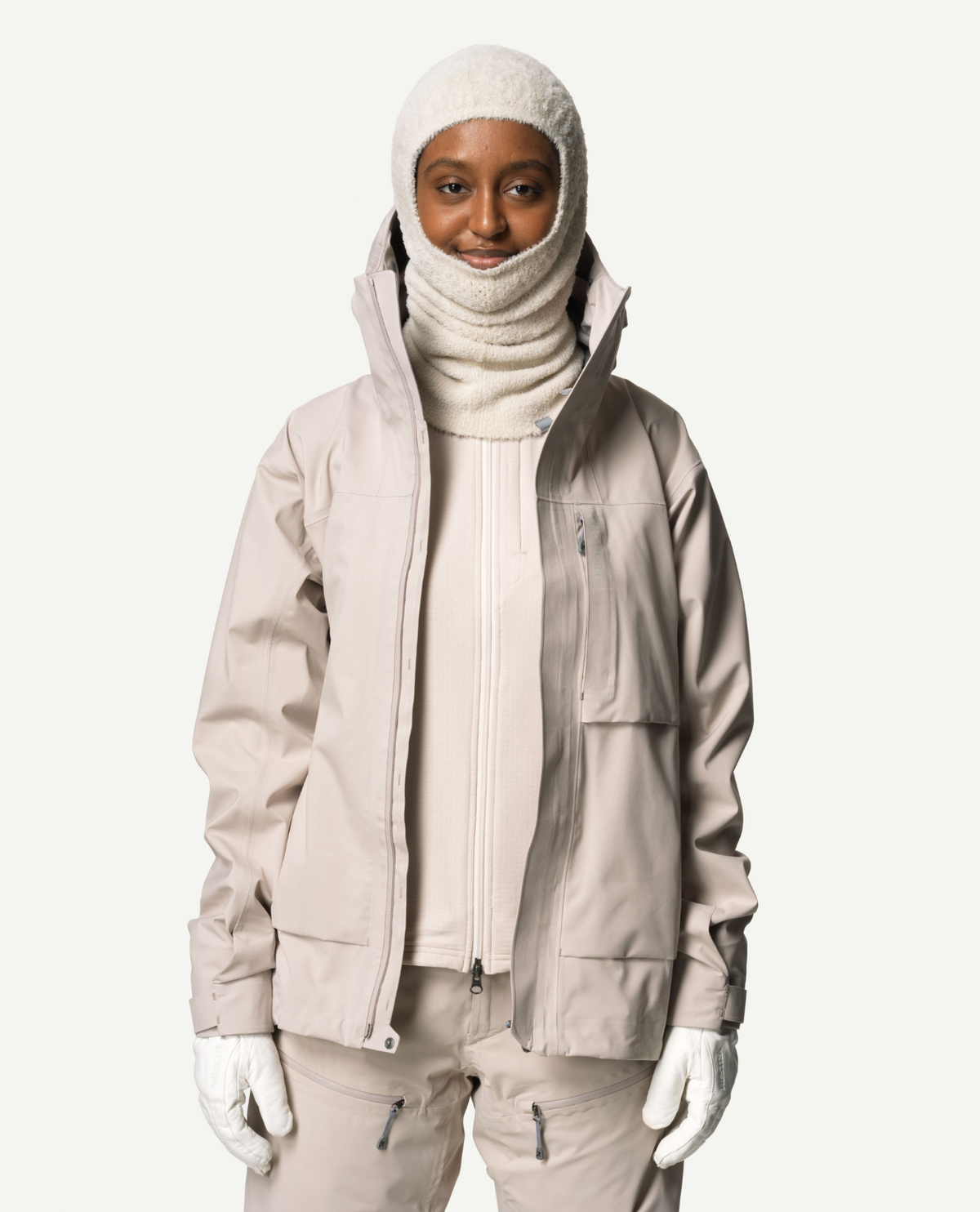 En person bär Houdini Lykan Neck Gaiter i färgen Foggy Mountain tillsammans med matchande beige skidjacka och vita handskar.