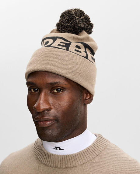 J.Lindeberg Tilo Beanie i brun färg med tofs buren av en modell mot vit bakgrund.