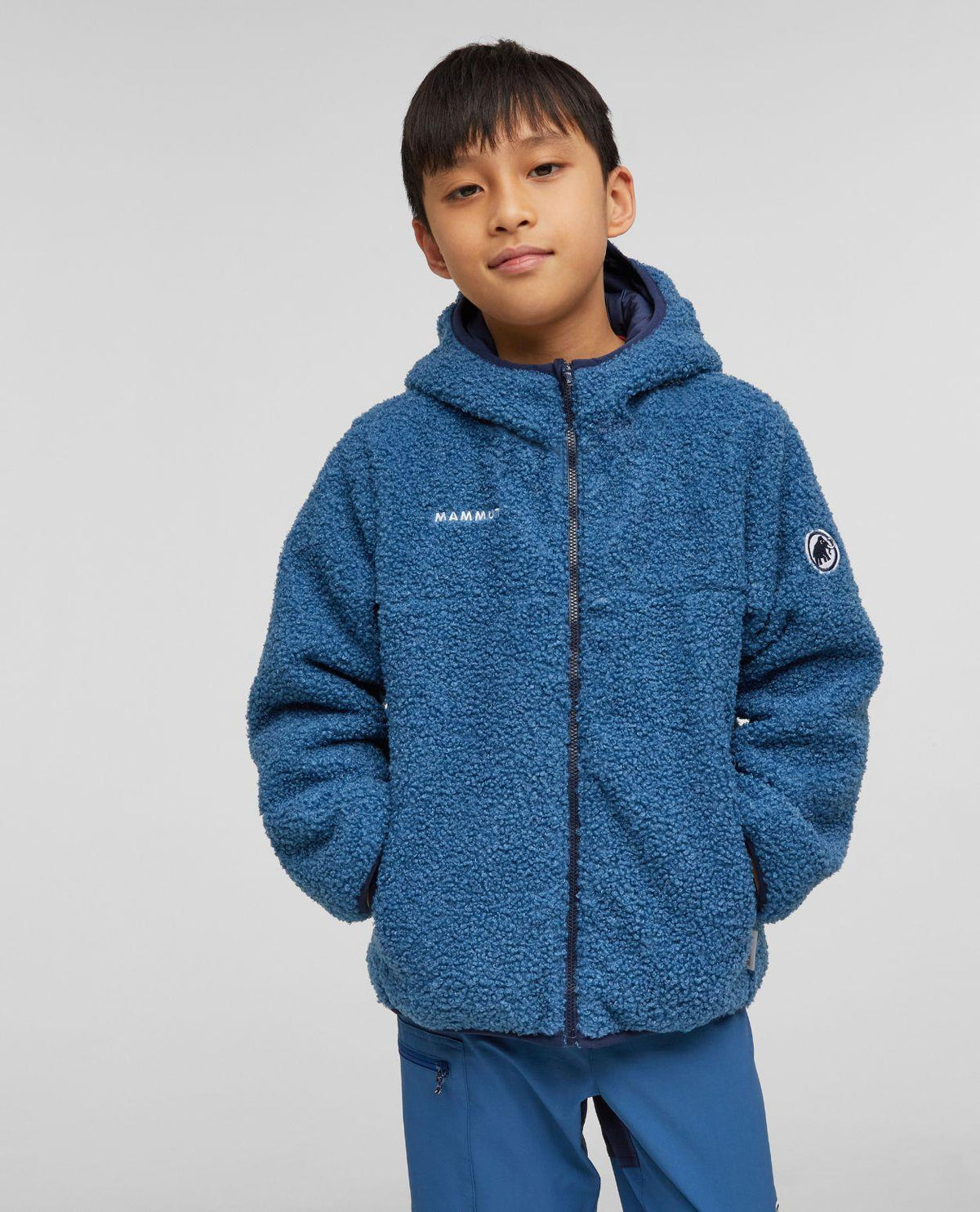Mammut Waymarker IN Reversible Hooded Jacket Junior i mörkblå teddyfleece buren av en ung skidåkare.