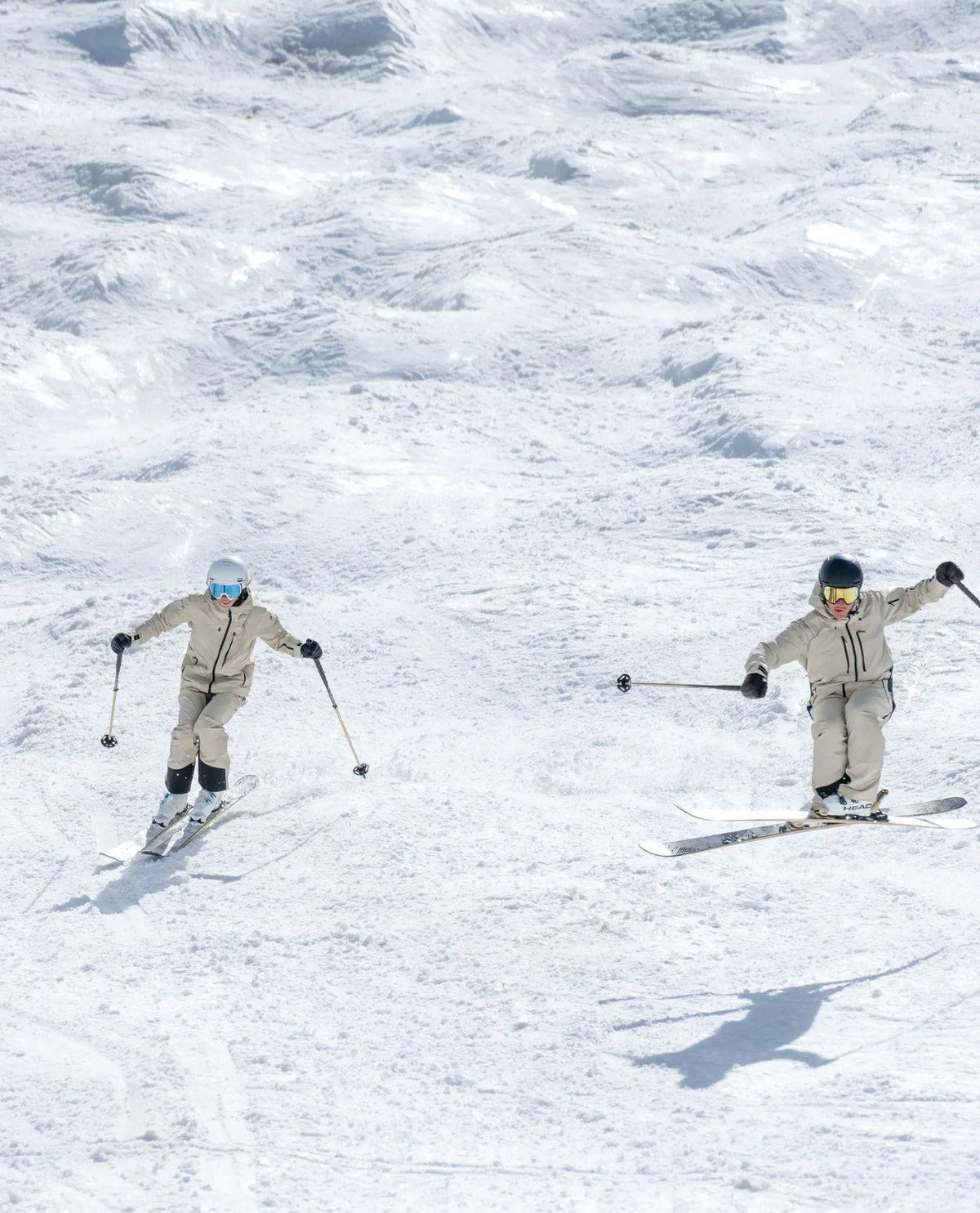 Två skidåkare i matchande ljusa kläder som åker skidor och gör ett hopp i ett snötäckt fjälllandskap.