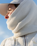 Person bär Houdini Lykan Neck Gaiter i beige färg runt halsen tillsammans med keps och jacka utomhus.