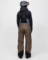 Bakifrånvy av dam i skidbyxor Peak Performance Vertical Gore-Tex Pro Bib Pants i färg terrain tan och ljusgrå med matchande jacka och hjälm.