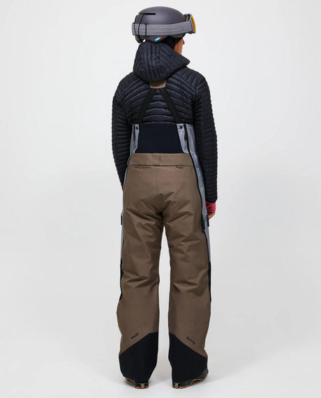 Bakifrånvy av dam i skidbyxor Peak Performance Vertical Gore-Tex Pro Bib Pants i färg terrain tan och ljusgrå med matchande jacka och hjälm.
