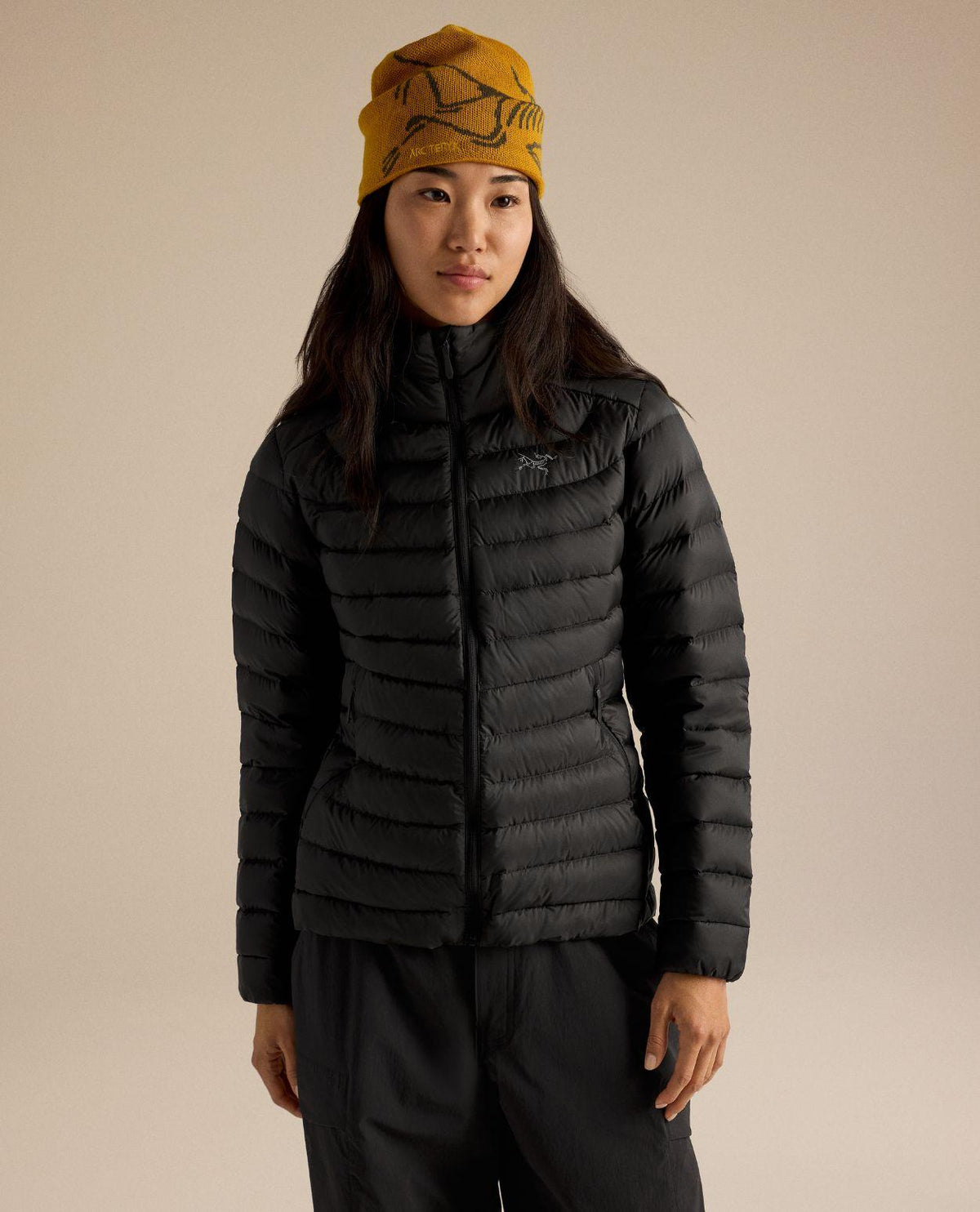 Kvinna i svart Arc'teryx Cerium Hoody lättviktsjacka med dunisolering och gul mössa.