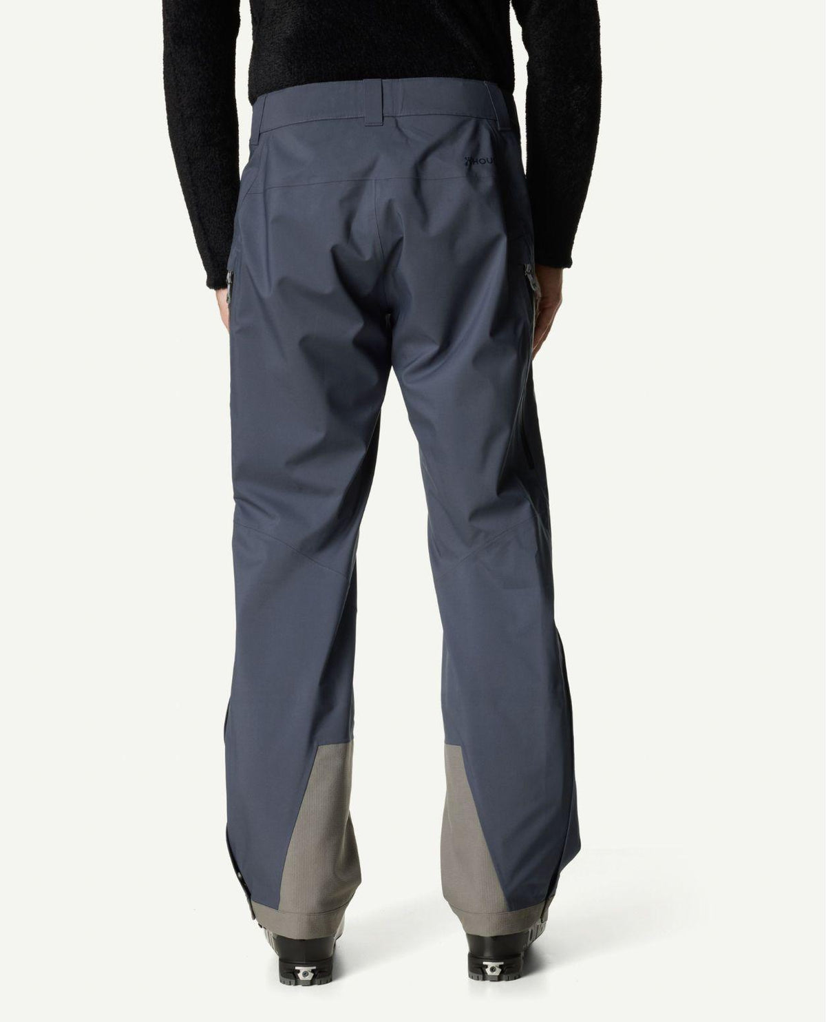 Houdini Ride Pants II för herrar i blå färg visas bakifrån på en skidåkare med pjäxor.