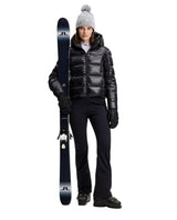 J.Lindeberg Nerina Down Jacket Women i svart, visad med matchande skidbyxor, pjäxor och skidor.