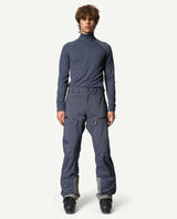 Manlig skidåkare iförd blå Houdini Ride Pants II skidbyxor och pjäxor framifrån.