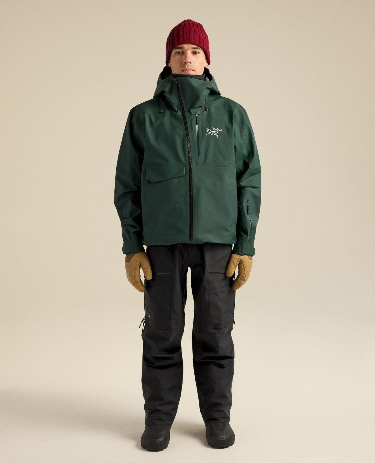 Arc'teryx Sabre Bib skidbyxor i svart för herr bärs här med grön skidjacka, röd mössa och bruna handskar.